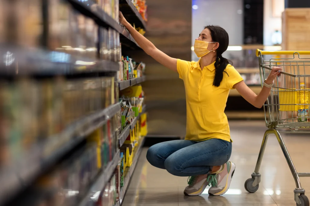 Woman wearing a mask shopping in a grocery store aisle, checking product labels and expiration dates using a shelf life calculator